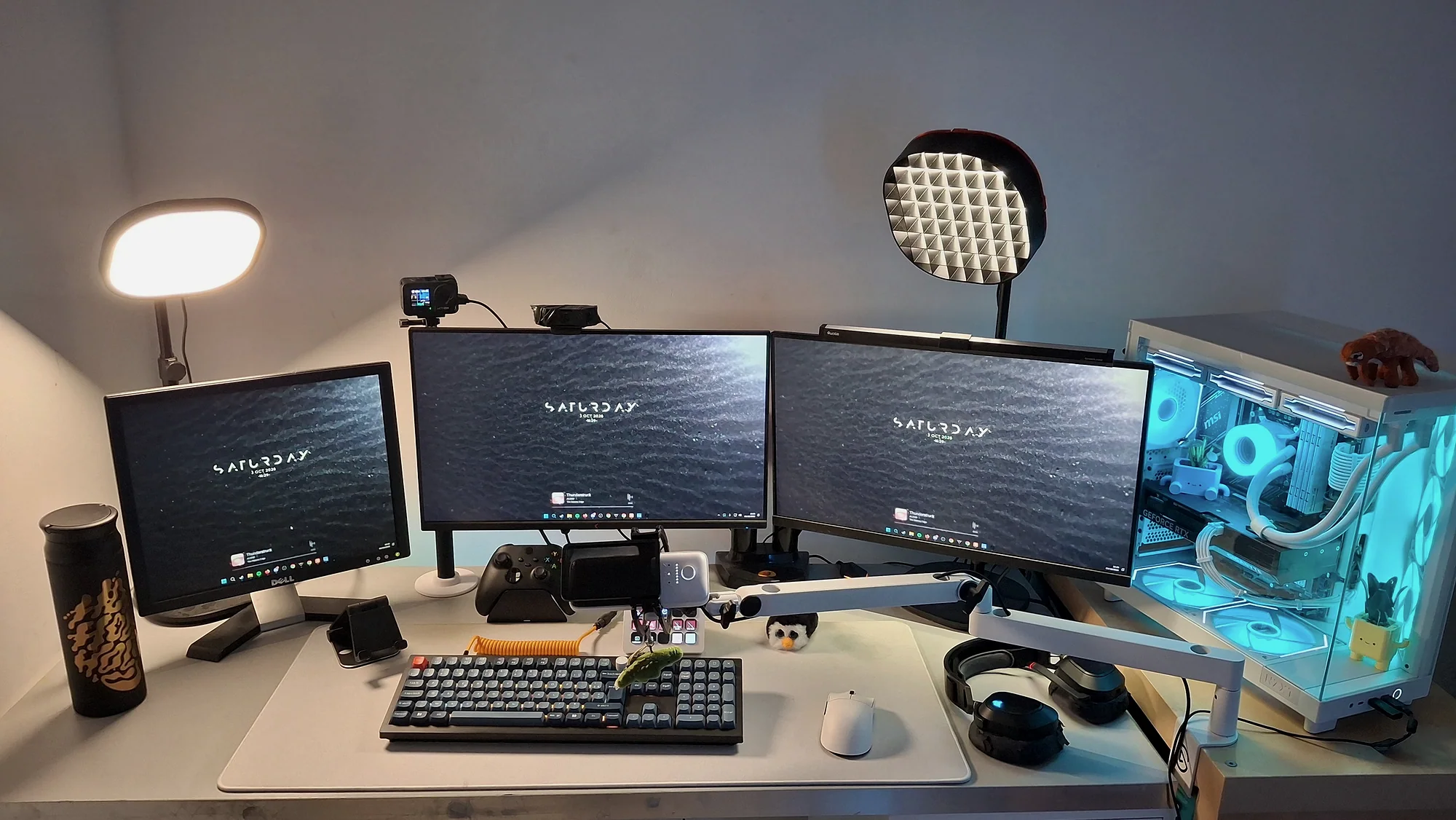 Vue d'ensemble du setup : trois écrans sur un bureau blanc, deux lumières de stream, le clavier, le Stream Deck et le PC à droite, avec ses ventilateurs éclairés en bleu clair.